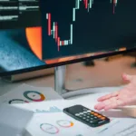 Pengusaha menganalisis data keuangan di monitor komputer dengan dokumen dan kalkulator di meja, mencerminkan proses evaluasi likuidasi perusahaan